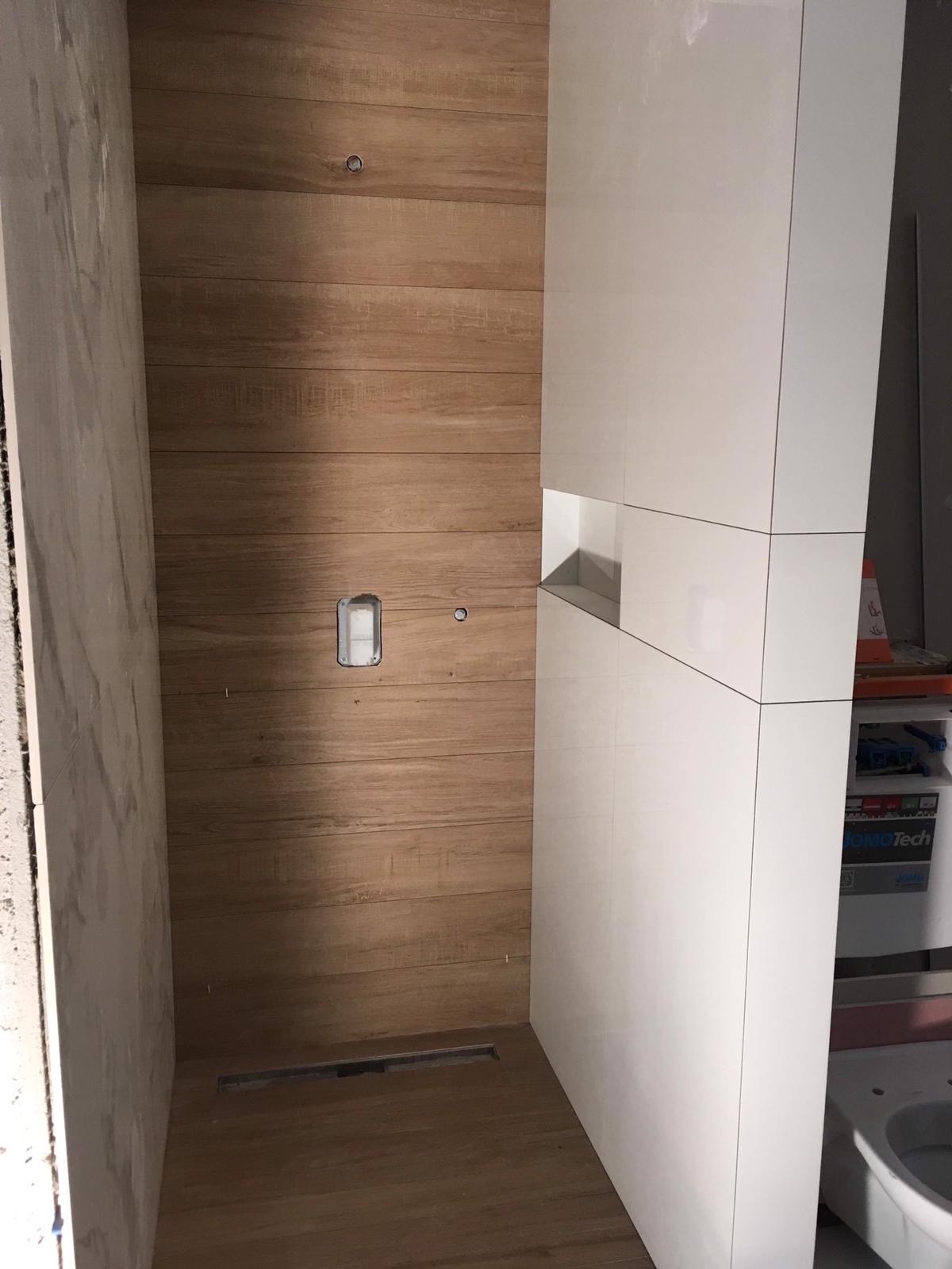 Wnętrze łazienki w trakcie wykończenia: drewnopodobne płytki na ścianie, biała zabudowa z wnęką, odpływ liniowy w podłodze, widoczna instalacja elektryczna.