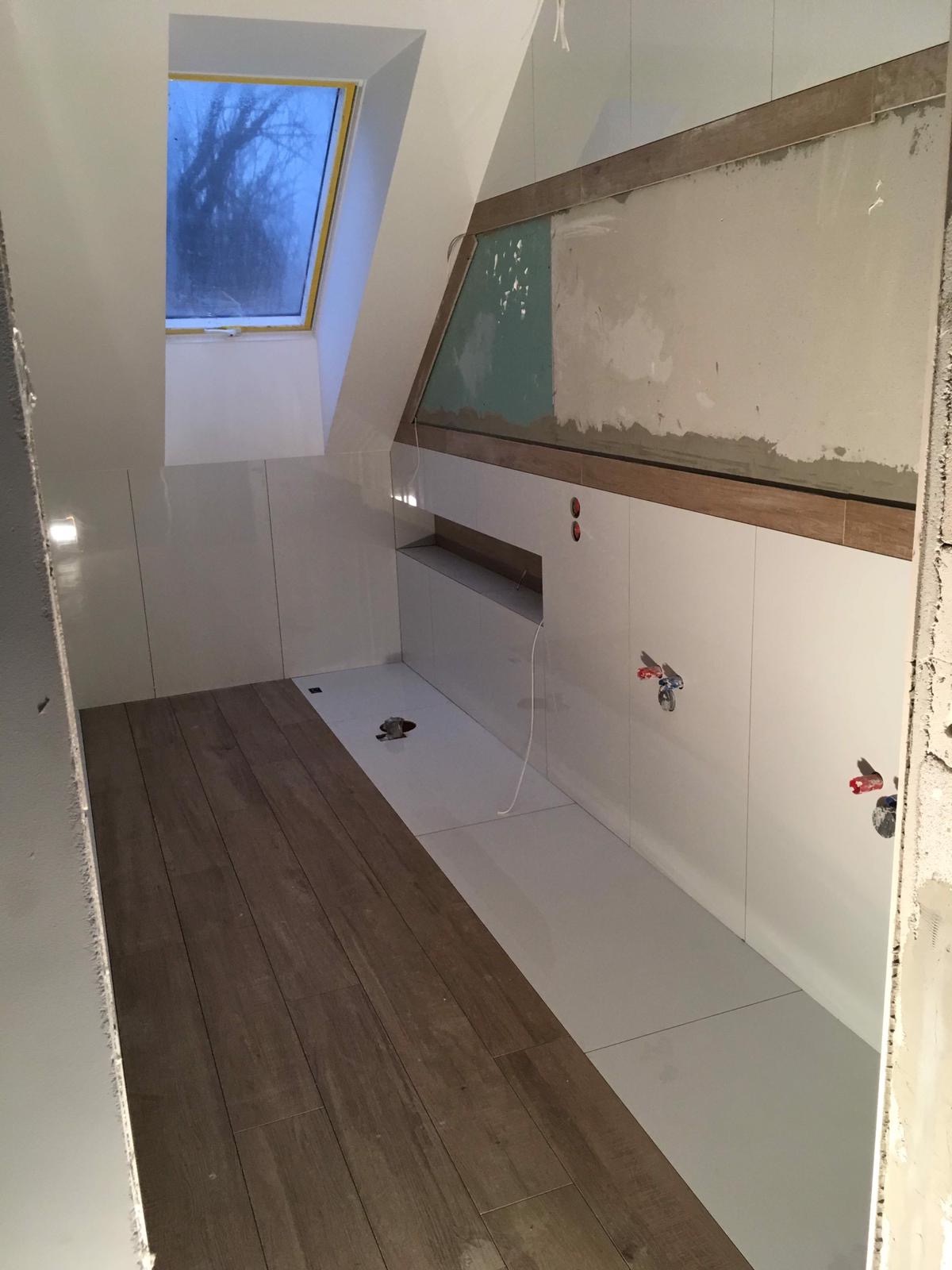 Układanie płytek w łazience na poddaszu: brązowe panele podłogowe przechodzą w białe płytki podłogowe, białe płytki ścienne z przygotowanymi otworami na armaturę, okno dachowe z widokiem na drzewa.