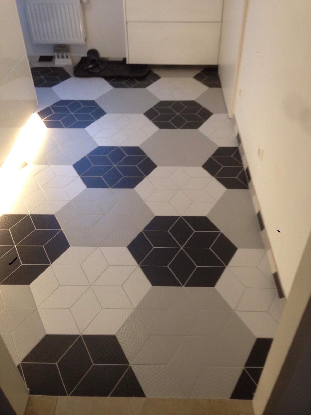 Ułożona podłoga z heksagonalnych płytek w odcieniach szarości, bieli i czerni, tworzących iluzję trójwymiarowych kostek; widoczny fragment grzejnika i szafki w tle.
