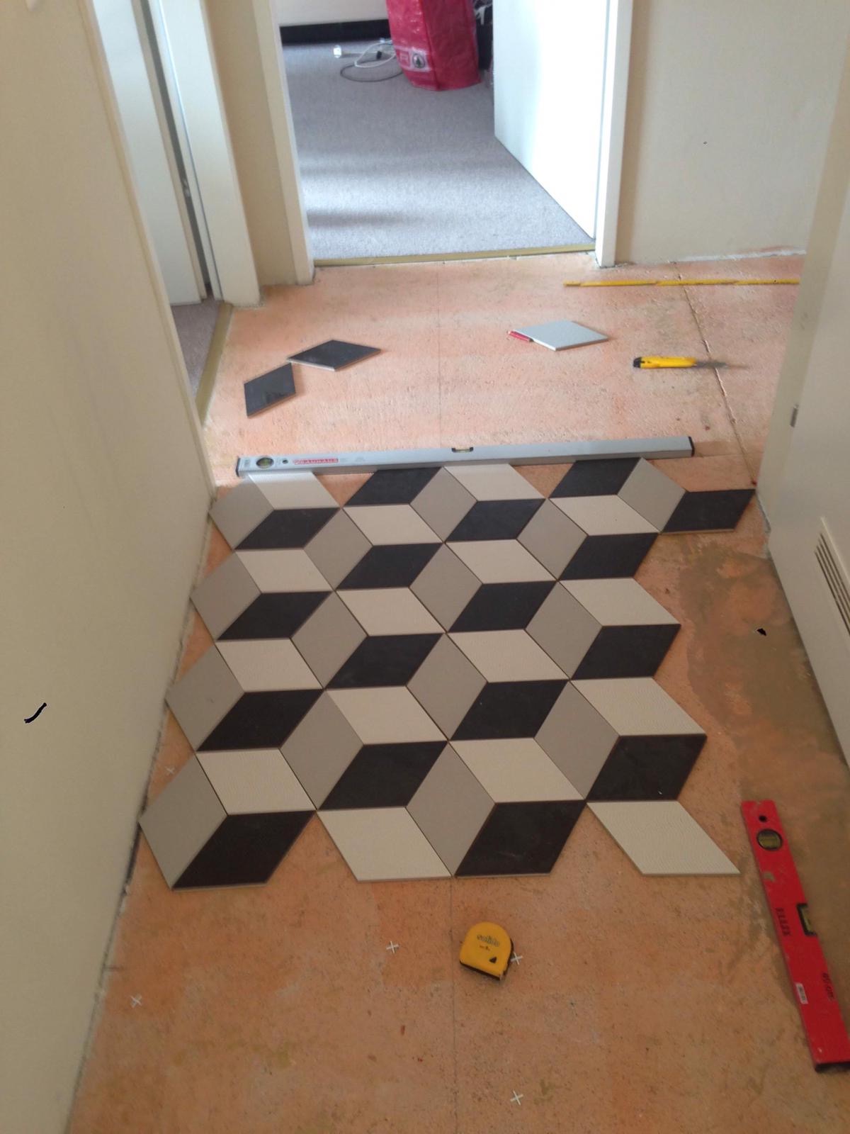 Układanie podłogi z płytek w geometryczny wzór 3D, widoczne narzędzia: poziomica, miarka, nóż do cięcia płytek.