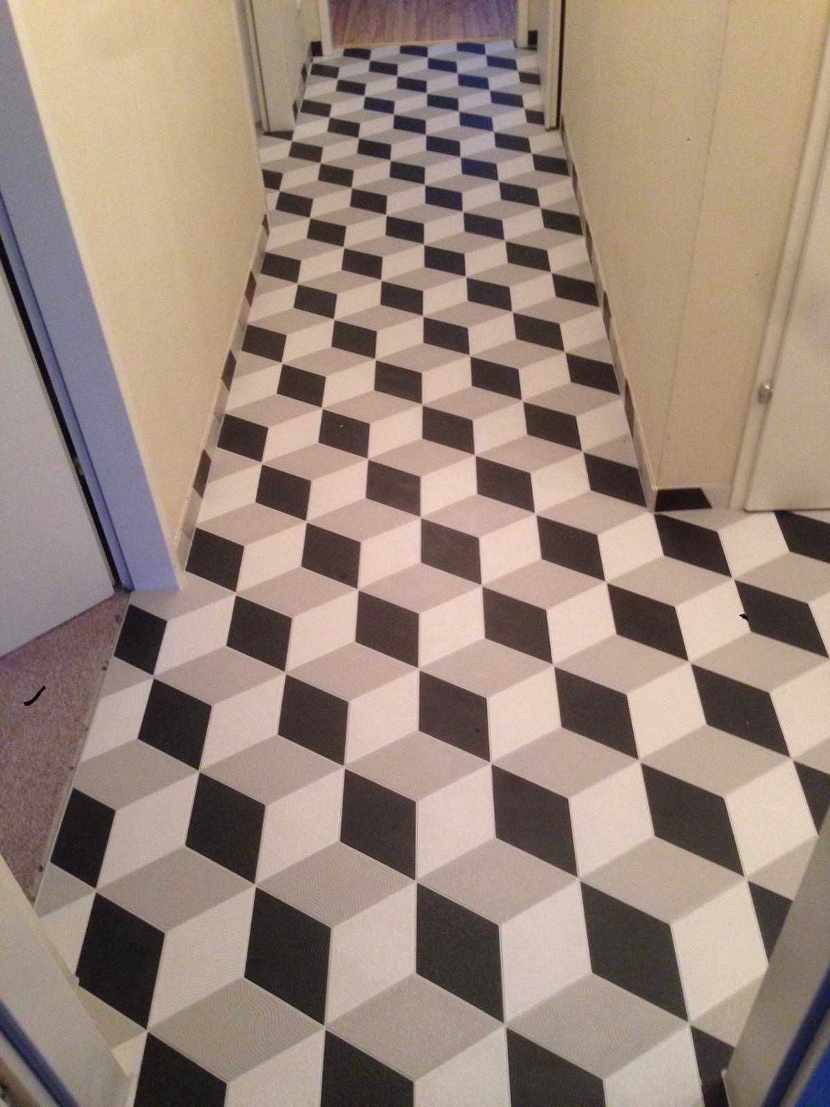 Korytarz z podłogą wyłożoną płytkami imitującymi trójwymiarowe kostki w odcieniach szarości i bieli, widoczne drzwi po bokach.