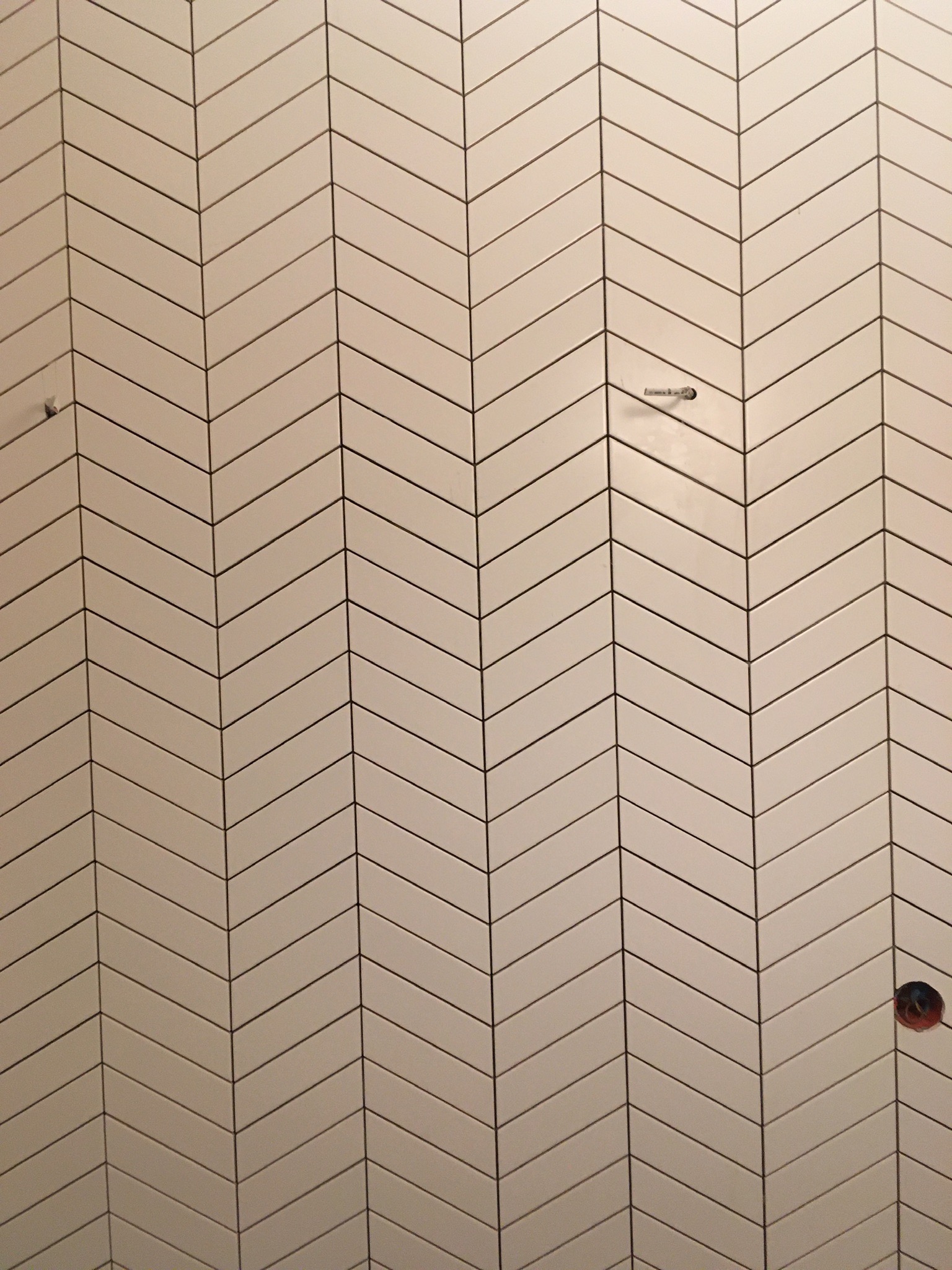 Ściana wyłożona białą, podłużną glazurą w jodełkę, z widocznymi otworami na gniazdka elektryczne i niedokładnościami fugowania.