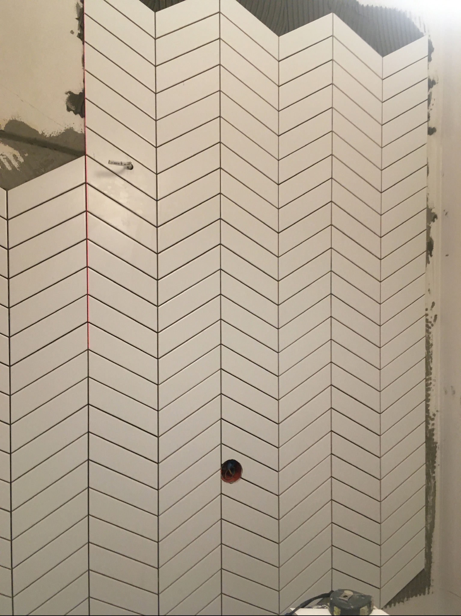 Ściana łazienki wyłożona białymi, podłużnymi płytkami ułożonymi we wzór jodełki, z widocznymi punktami instalacji wodnej i niewielkimi niedociągnięciami przy krawędziach.