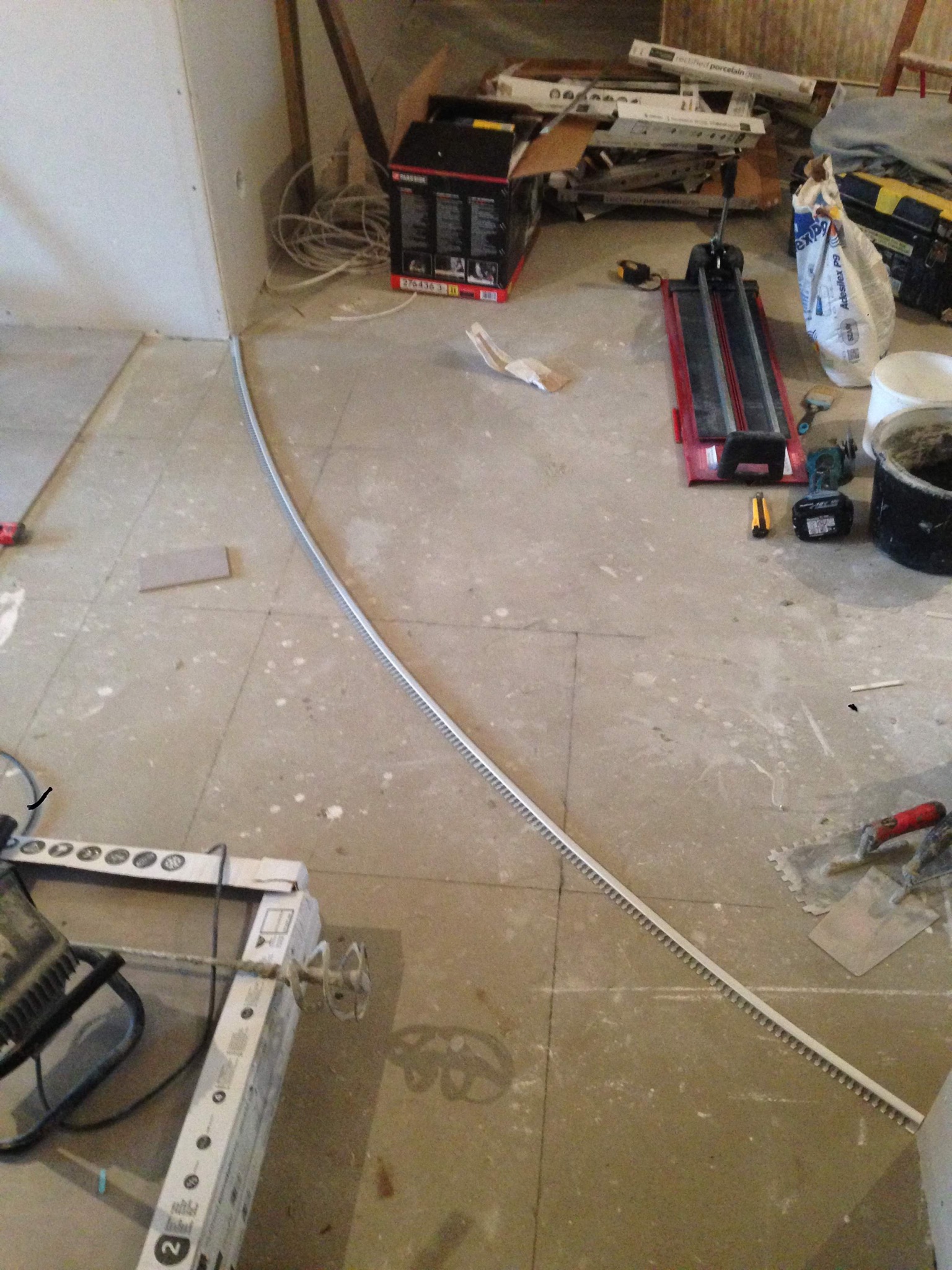 Przygotowanie do układania płytek: profil wykończeniowy łukowy, narzędzia do cięcia, mieszarka do zaprawy i wiadro z zaprawą na podłodze pokrytej płytkami w trakcie remontu.