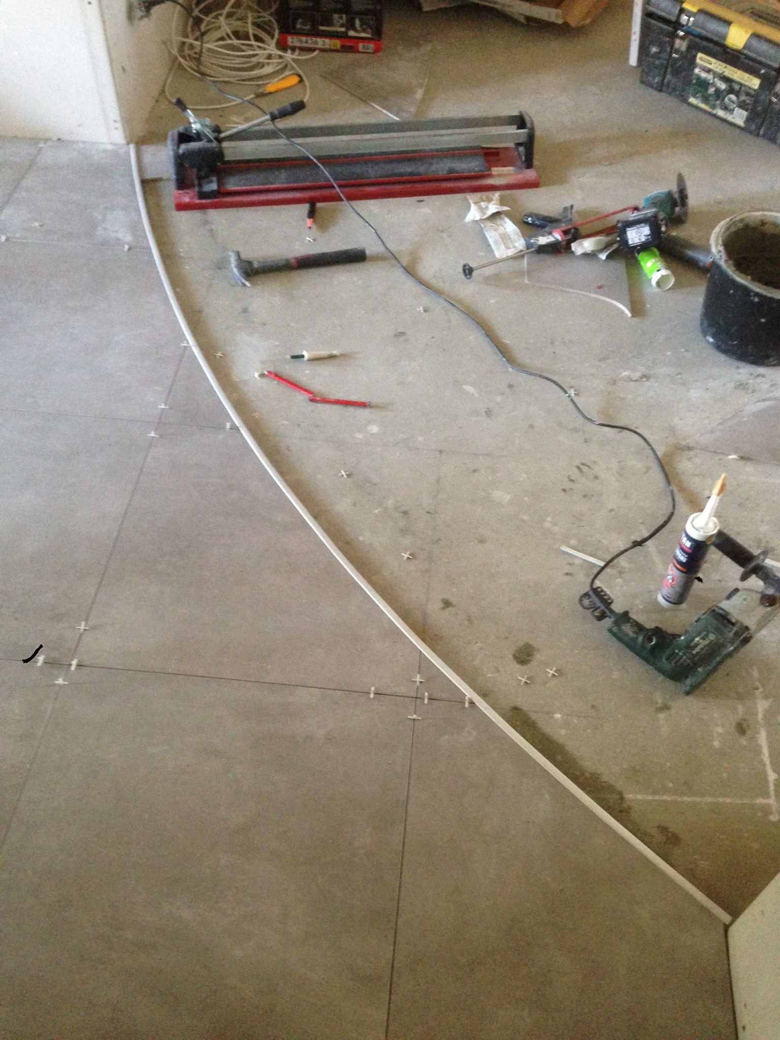 Układanie szarych płytek podłogowych z krzyżykami dystansowymi, widoczna listwa wykończeniowa, narzędzia glazurnicze: przecinarka do płytek, młotek, poziomica, wiertarka z klejem