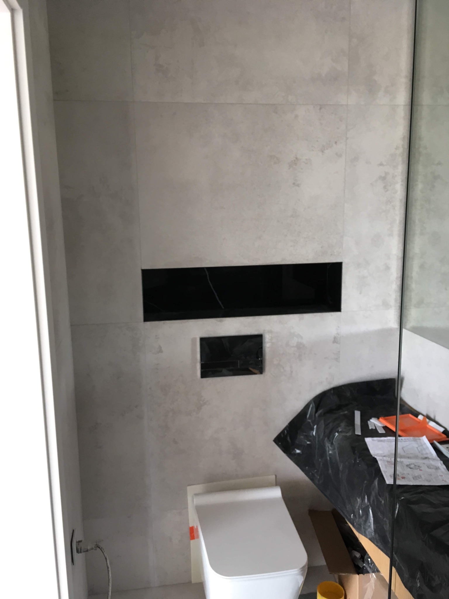 Płytki w odcieniach szarości z czarną wnęką i zabudową toalety w trakcie montażu, widoczne narzędzia i instrukcje na czarnej folii ochronnej.