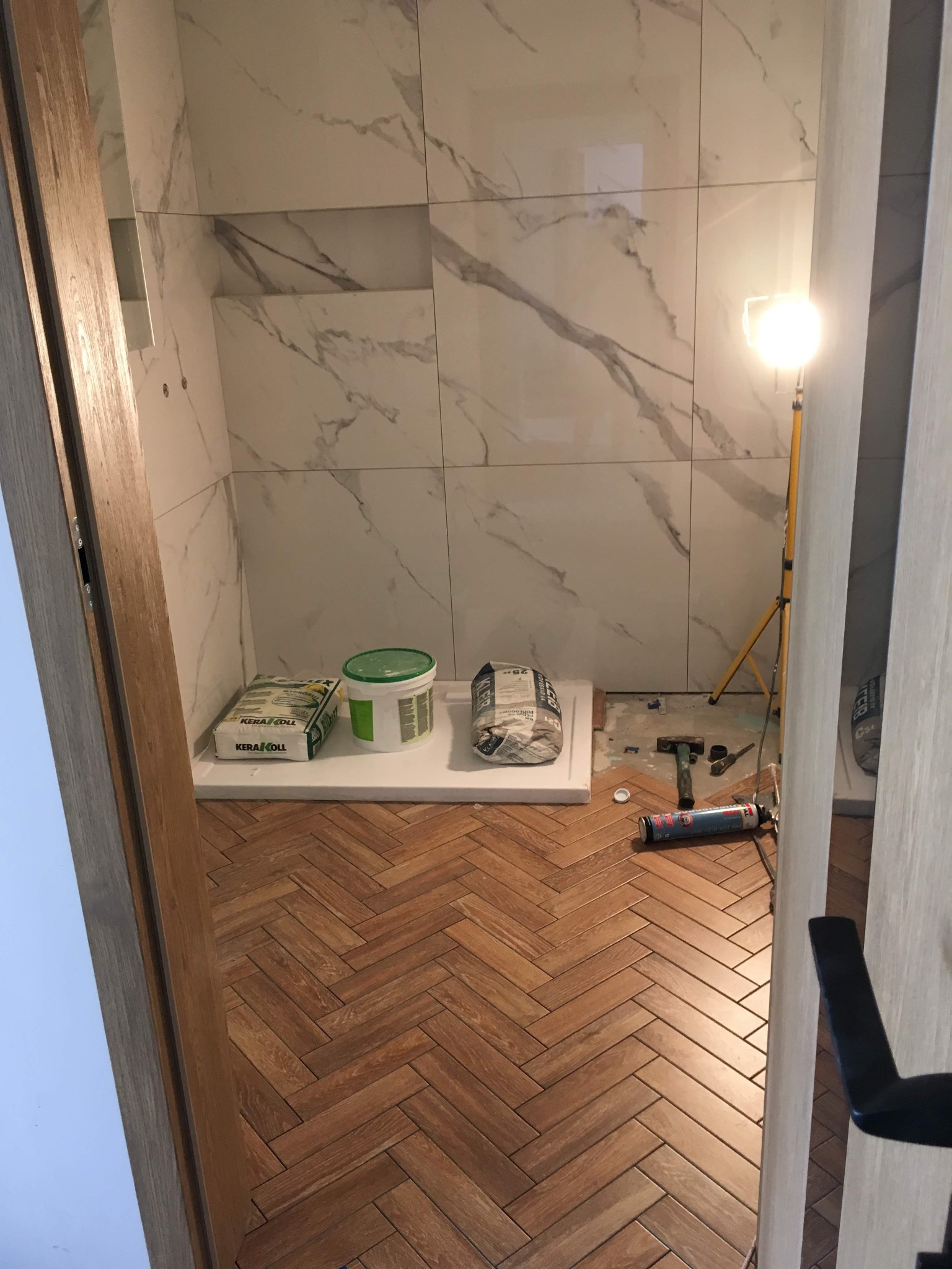 Montaż płytek imitujących marmur w łazience, widoczne narzędzia, klej i fugi na podłodze z parkietu w jodełkę, oświetlenie halogenowe.