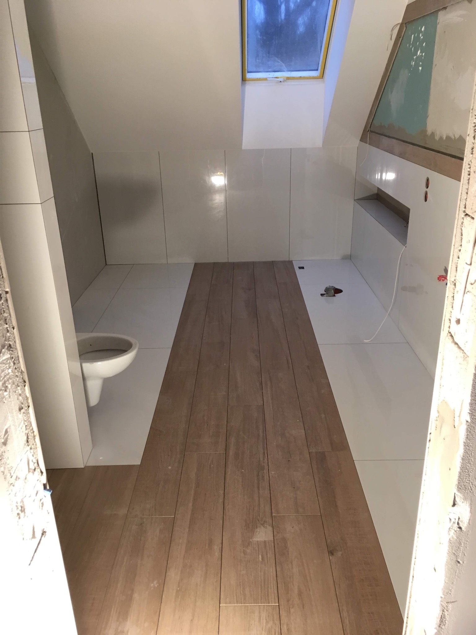 Łazienka na poddaszu w trakcie wykańczania: białe płytki ścienne, drewnopodobne płytki podłogowe ułożone częściowo, widoczny fragment toalety i okno dachowe.