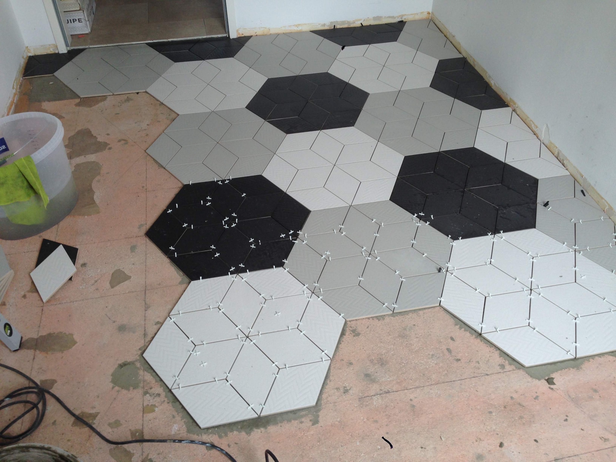 Układanie podłogi z heksagonalnych płytek w odcieniach szarości i czerni, z użyciem krzyżyków dystansowych, widoczny poziomica i wiadro z zaprawą.