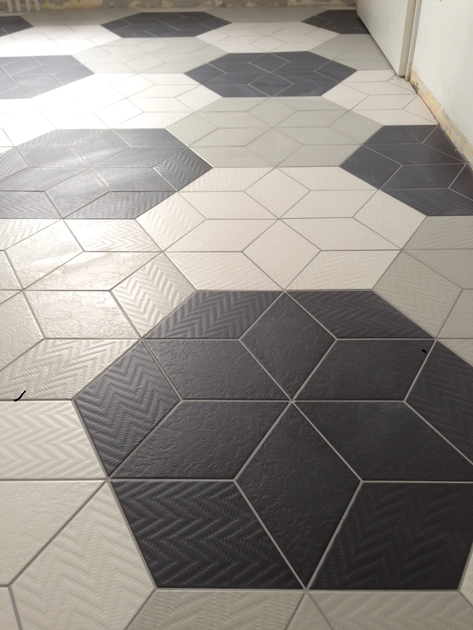 Wzorzysta podłoga z heksagonalnych płytek w odcieniach szarości i bieli, ułożonych w geometryczny wzór 3D, fuga w kolorze jasnoszarym, widoczny fragment ściany w tle.