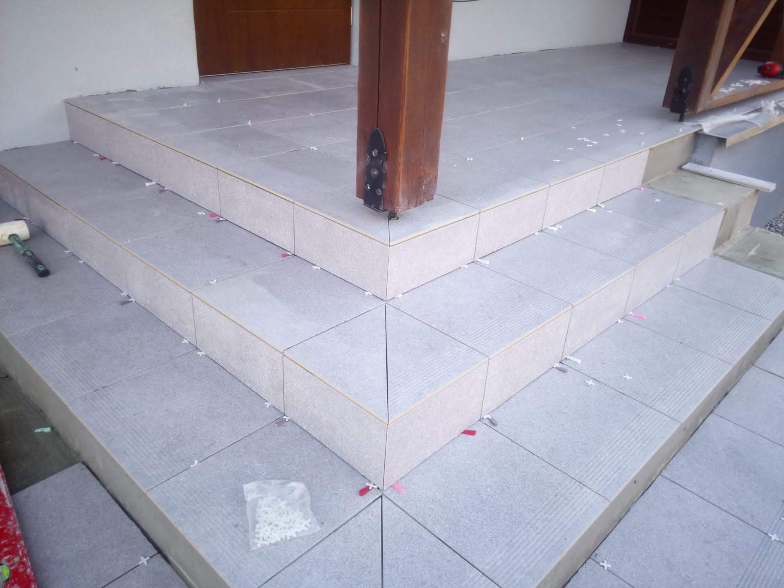 Wyłożone szarymi płytkami schody wejściowe, widoczne krzyżyki dystansowe, gumowy młotek i paczka krzyżyków, drewniany słup z metalowymi okuciami.