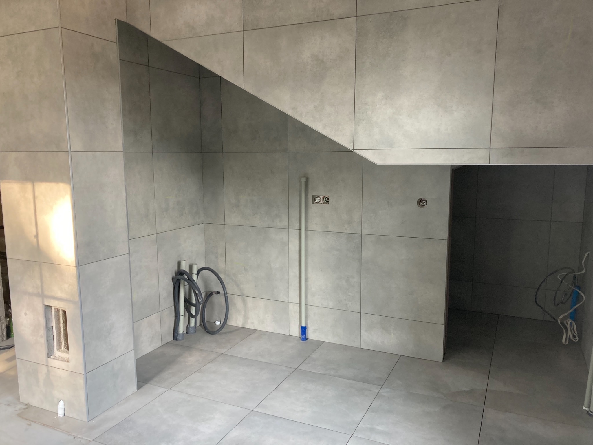 Szare, matowe płytki ceramiczne ułożone na ścianach i podłodze w pomieszczeniu w trakcie remontu, widoczne rury i kable instalacyjne, surowe wykończenie otworu w ścianie.