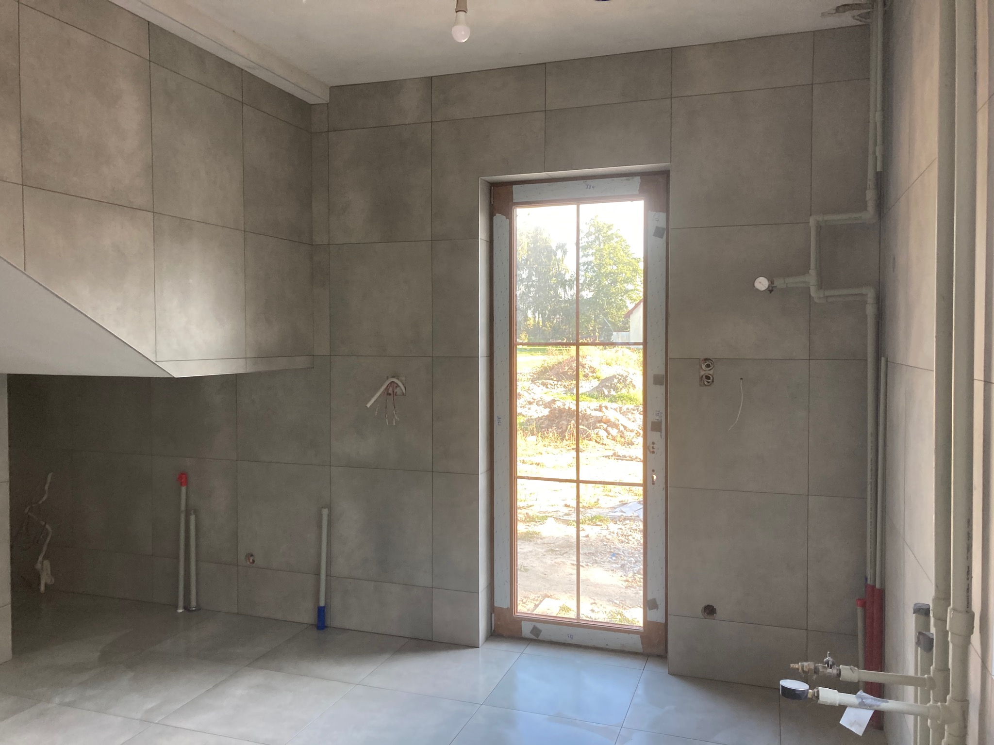 Szare, matowe płytki ceramiczne ułożone na ścianach i podłodze w pomieszczeniu z oknem, widoczne wystające rury instalacji wodnej i elektrycznej, surowy stan wykończenia.