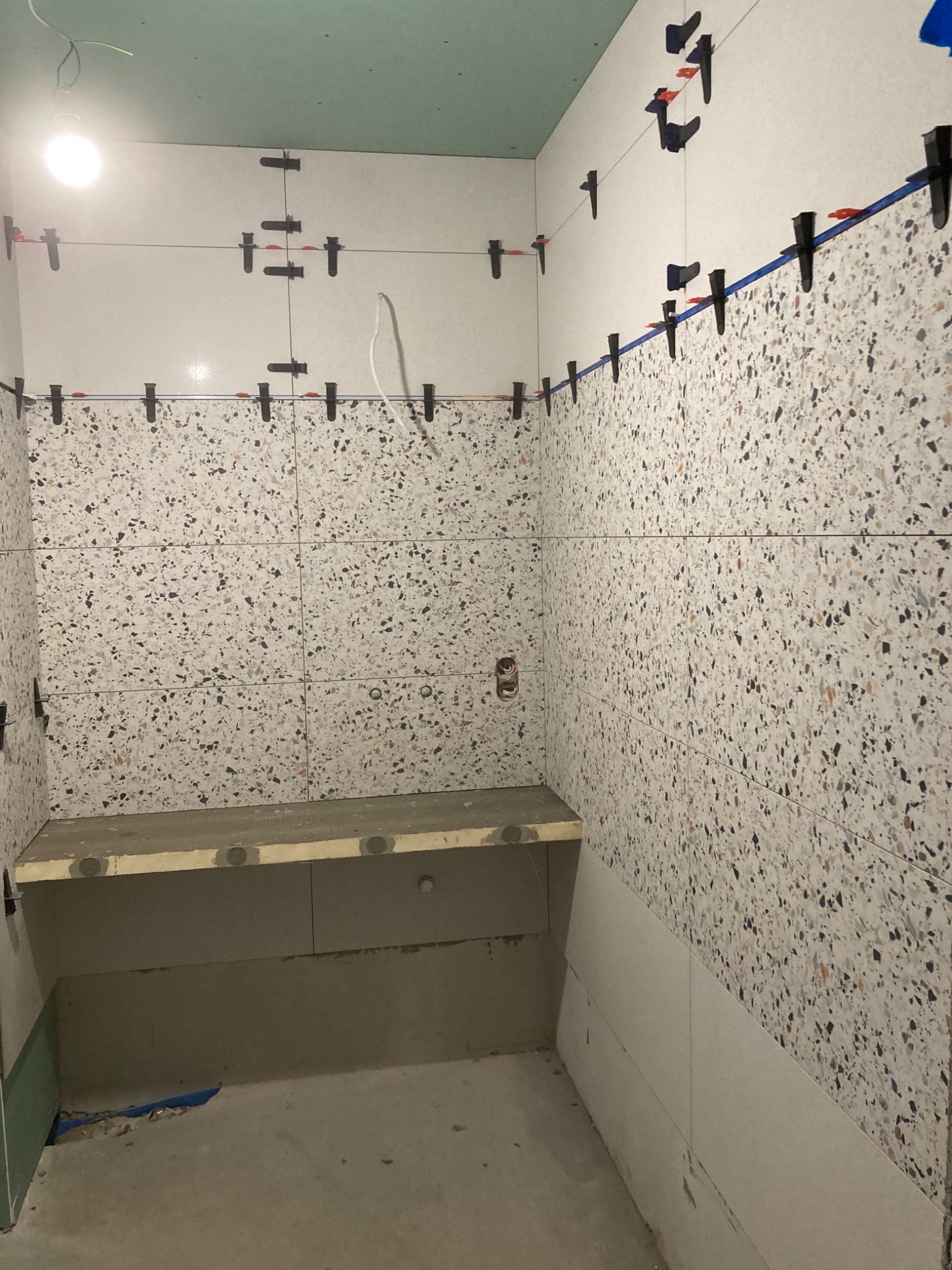 Ściany łazienki w trakcie wykańczania, z widocznymi płytkami imitującymi lastryko, system poziomowania płytek z czarnymi klipsami i czerwonymi klinami, oraz surowy betonowy stelaż pod umywalkę.