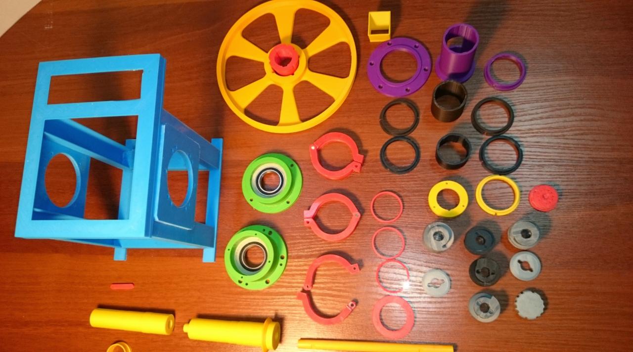 Różnokolorowe, plastikowe elementy, prawdopodobnie wydrukowane w 3D, ułożone na drewnianym blacie. Widoczne są m.in. niebieska rama z otworami, żółte koło zębate, zielone i czerwone pierścienie...