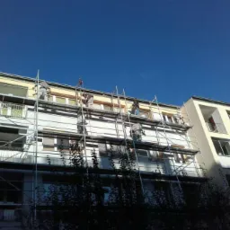 Pracownicy na rusztowaniu malują elewację bloku mieszkalnego na tle błękitnego nieba. Widoczne drzewa w dolnej części kadru.