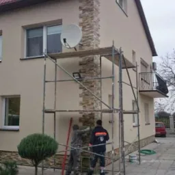 Prace wykończeniowe elewacji domu z użyciem kamienia dekoracyjnego. Dwóch pracowników na rusztowaniu, elewacja w kolorze beżowym, czerwony dach.