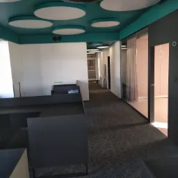 Wnętrze biura w trakcie wykańczania: szara wykładzina dywanowa, nowoczesne biurka w kolorze grafitowym, przeszklone ściany działowe z widokiem na jasne pomieszczenia, sufit w odcieniach turkusu...