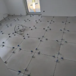 Ułożone płytki podłogowe w jasnym pomieszczeniu z białymi ścianami, użyto systemu poziomowania z niebieskimi klipsami i czarnymi krzyżykami, widoczne przewody na podłodze.