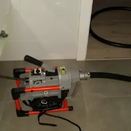 Elektryczna przepychaczka do rur z czarnym wężem i spiralą roboczą, stojąca na szarych płytkach pod otwartą szafką.