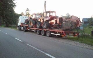 Ciężarówka z naczepą niskopodwoziową przewożąca dwa traktory rolnicze na tle wiejskiego krajobrazu.