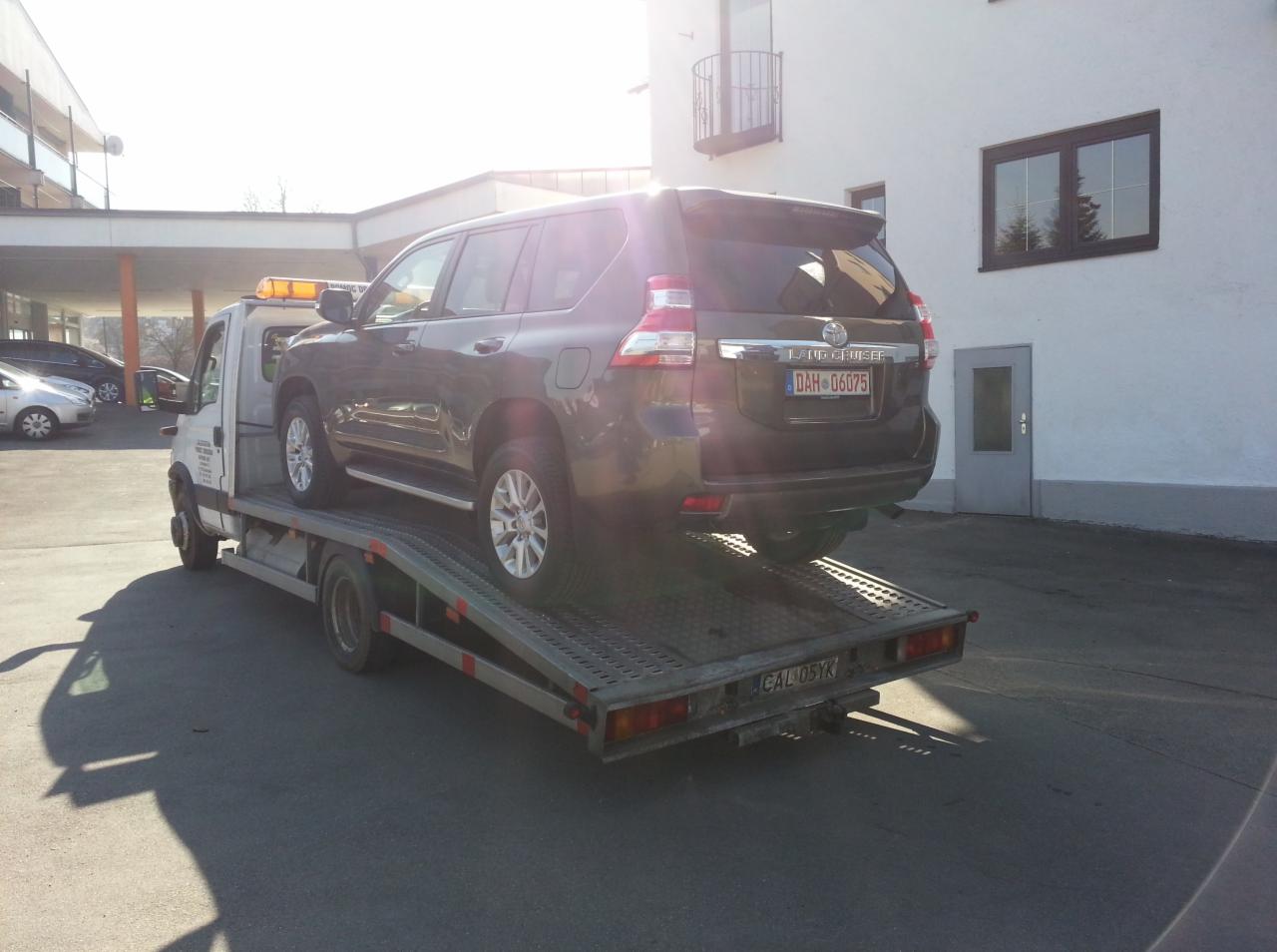 Szary samochód terenowy Toyota Land Cruiser na lawecie podczas słonecznego dnia, widoczne tablice rejestracyjne DAH-06075 oraz CAL 05YK, budynek w tle.