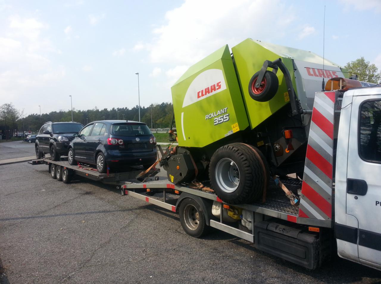 Laweta przewozi dwa samochody osobowe oraz prasę rolniczą Claas Rollant 355, zabezpieczone pasami, na parkingu.