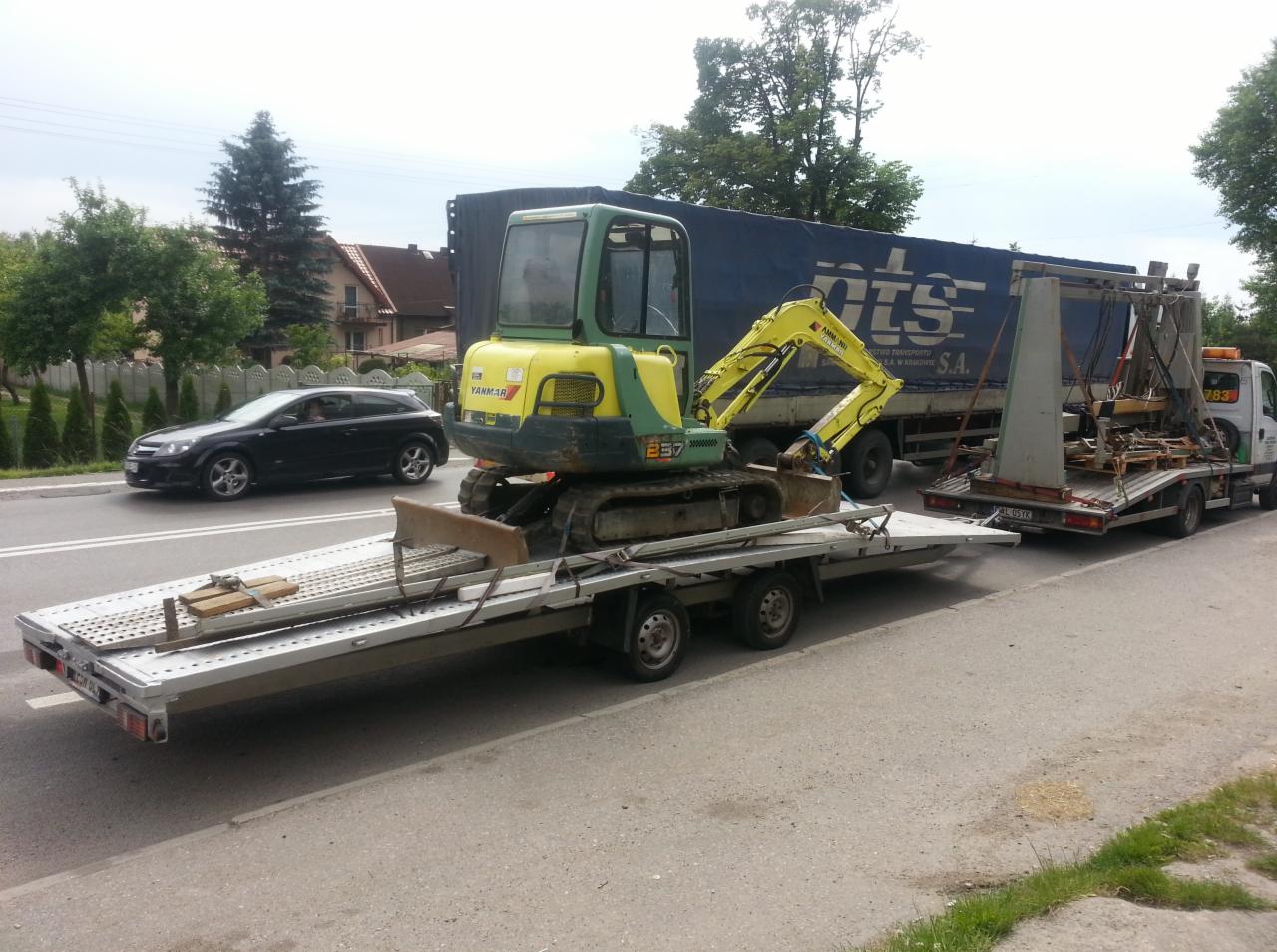 Żółto-zielona mini koparka Yanmar na niskopodwoziowej przyczepie lawety, za nią niebieska plandeka ciężarówki i druga laweta z ładunkiem, czarny samochód osobowy w tle.