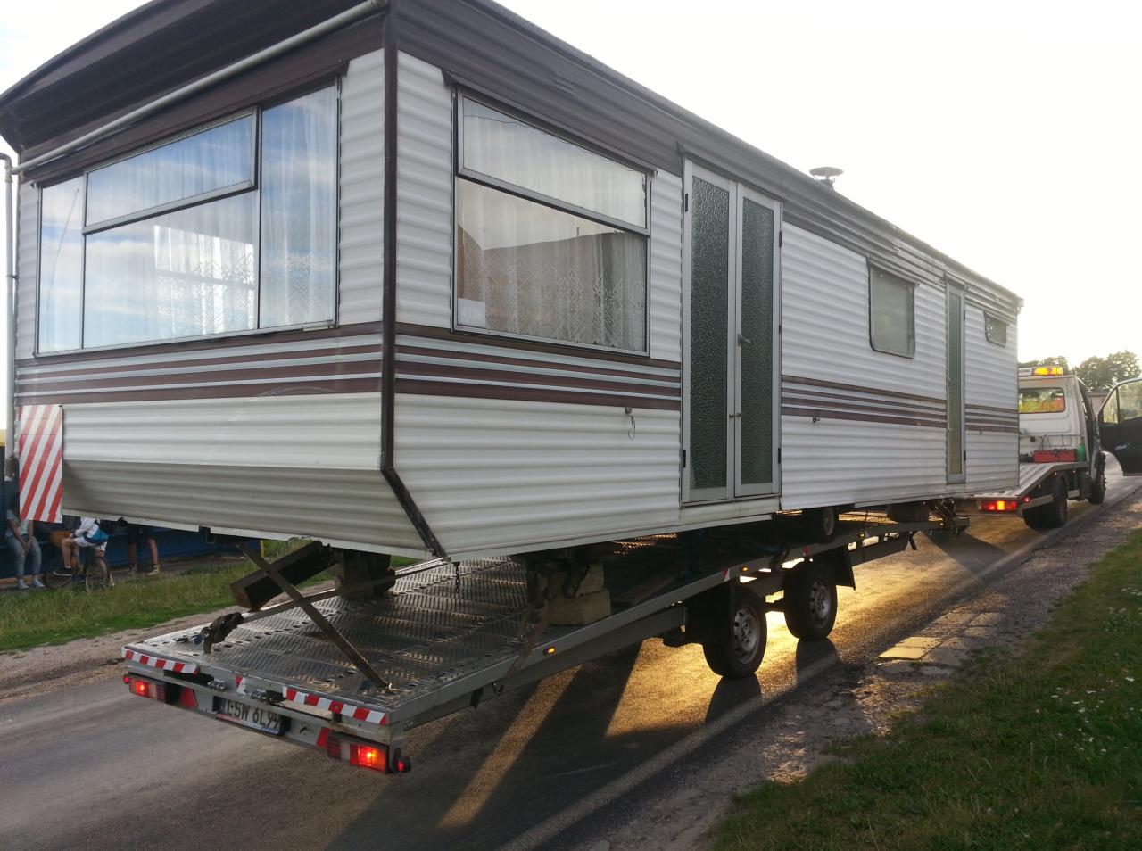Przyczepa kempingowa transportowana na niskopodwoziowej lawecie, widoczne światła lawety i fragment ciągnącego ją pojazdu, z boku widoczni ludzie.