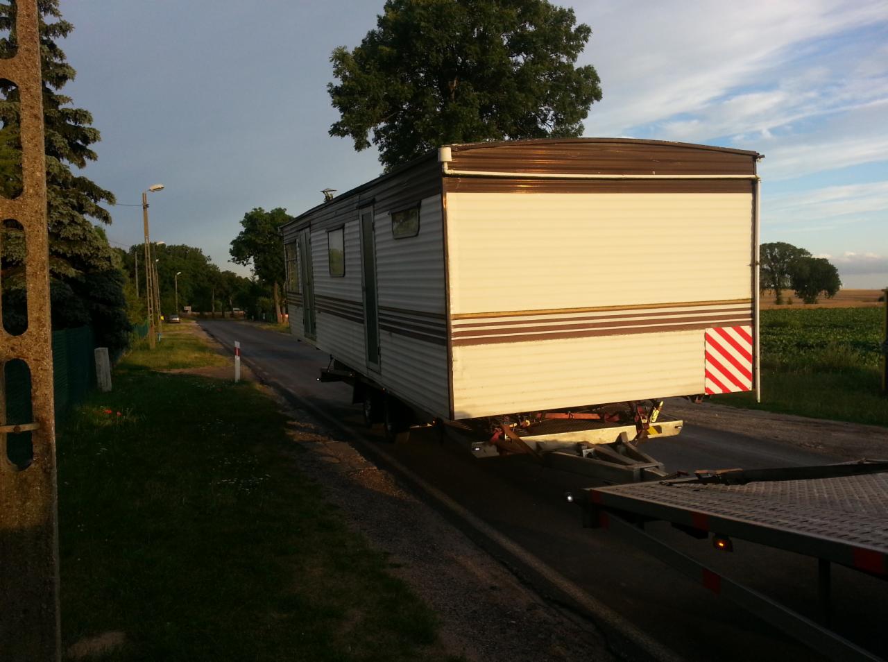 Przyczepa kempingowa przewożona na niskopodwoziowej lawecie na wąskiej asfaltowej drodze wiejskiej, widoczne drzewa i pole w tle.