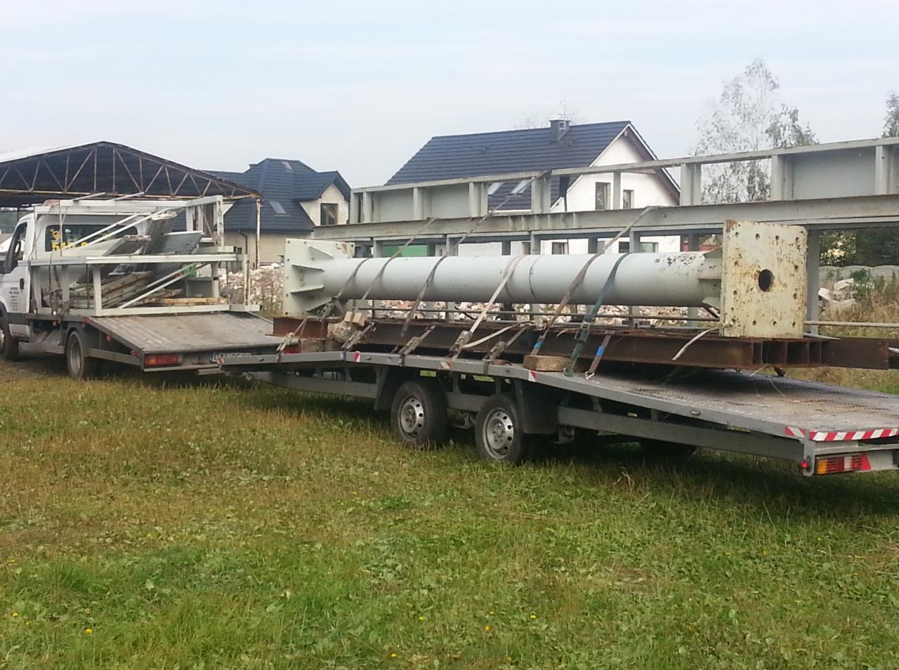 Laweta z ładunkiem w postaci metalowej rury z elementami mocującymi, Ciechocinek; na drugim planie zabudowania.