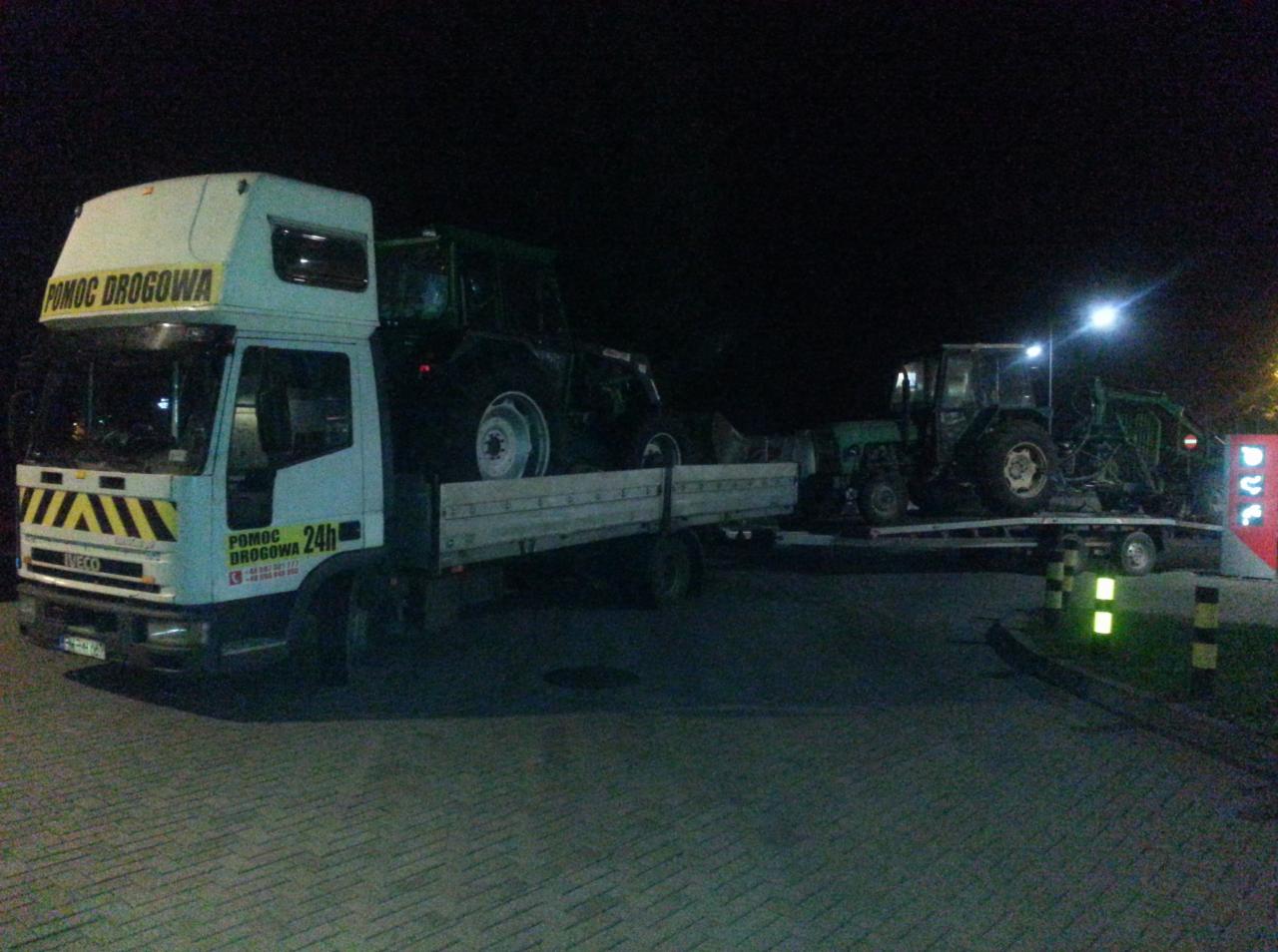 Biała ciężarówka pomocy drogowej z napisem 'POMOC DROGOWA 24h' przewozi dwa zielone traktory na platformie w nocy. Na dachu ciężarówki również widnieje napis 'POMOC DROGOWA'.