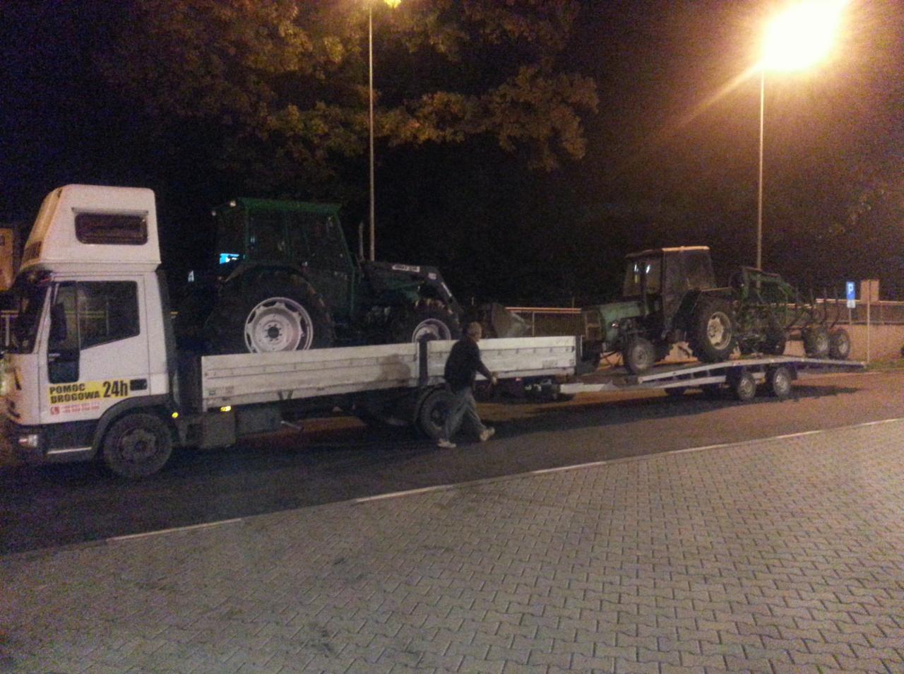 Laweta z dwoma traktorami, jeden zielony z przodu i mniejszy zielony z tyłu, na tle nocnego oświetlenia ulicznego, z widocznym napisem 'Pomoc Drogowa 24h' na kabinie.