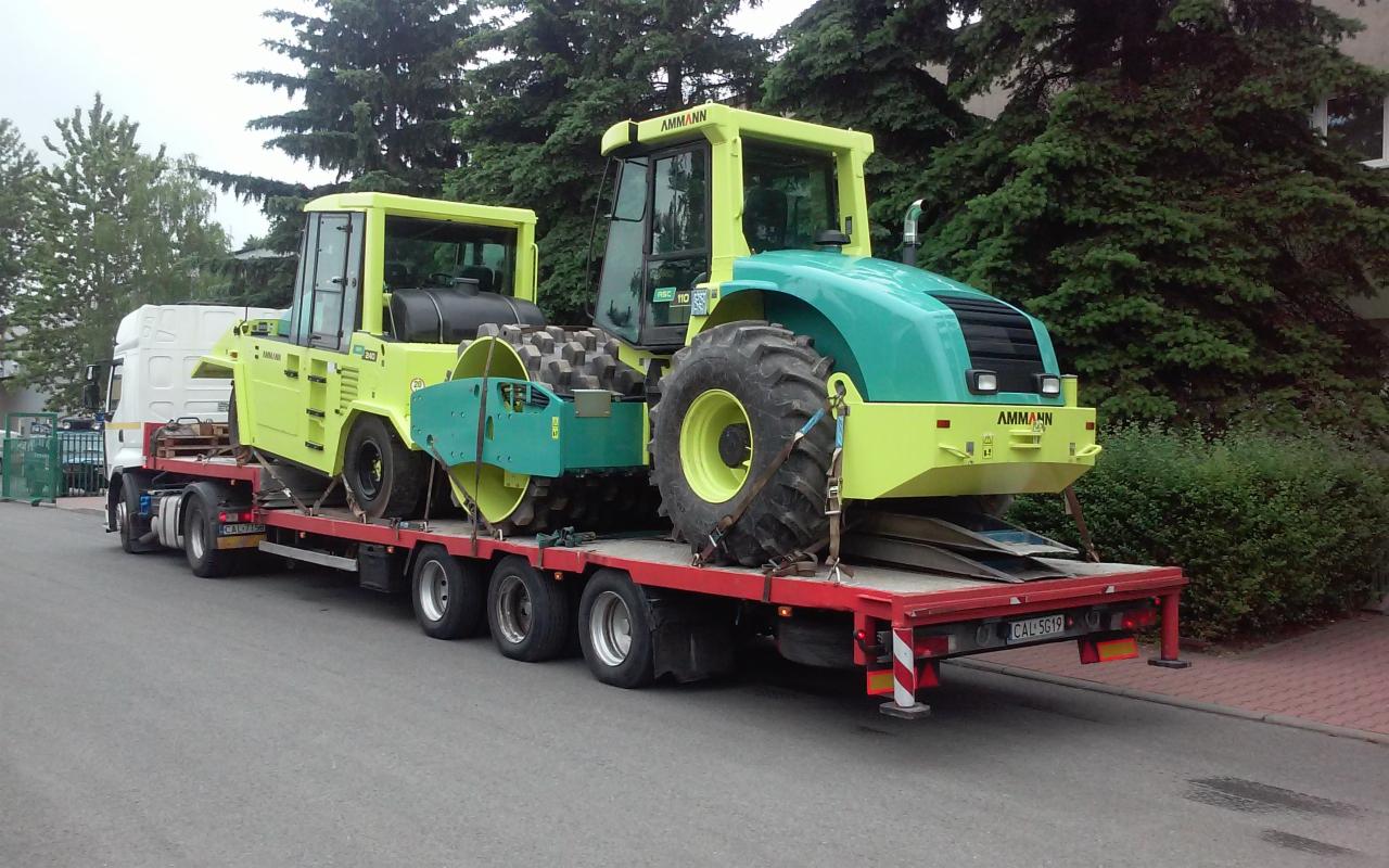 Dwa walce drogowe marki Ammann, jeden zielono-żółty z przodu i drugi żółty z tyłu, zabezpieczone pasami na czerwonej niskopodwoziowej naczepie ciężarówki na tle zielonych drzew.