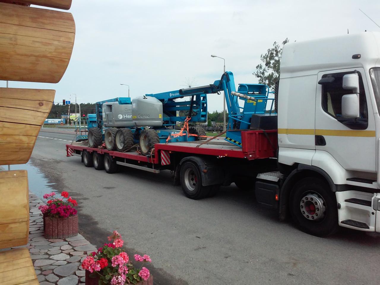 Ciężarówka z białą kabiną przewozi na czerwonej platformie dwie niebieskie podnośniki koszowe marki Heracle Genie. Widok z boku, na pierwszym planie donice z kwiatami.