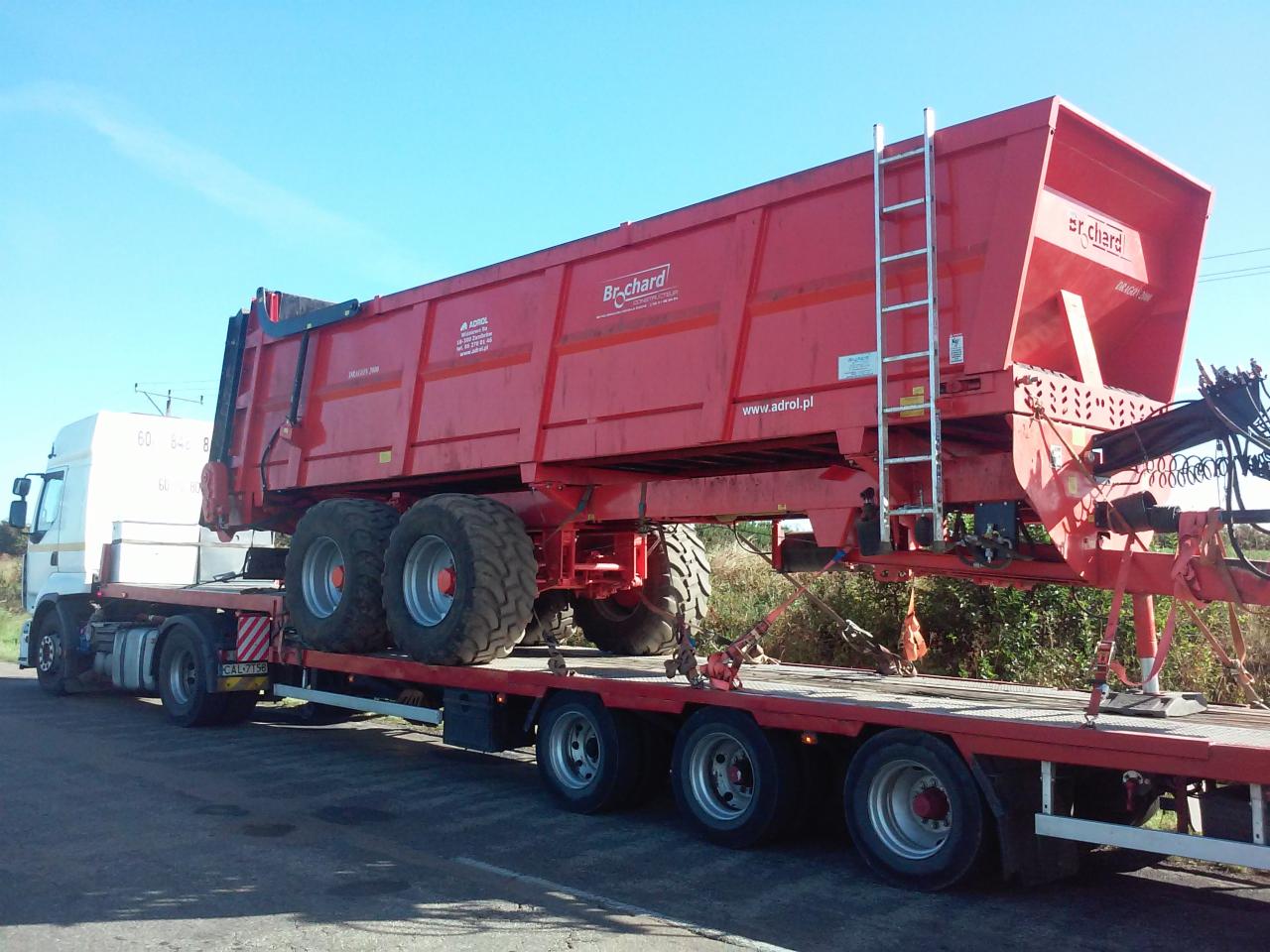 Ciężarówka z naczepą przewożąca dużą, czerwoną przyczepę rolniczą marki Br-chard z drabinką i napisem www.adrol.pl, widok z boku na tle błękitnego nieba.