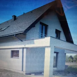 Nowoczesny dom jednorodzinny z ciemną dachówką, balkonem z metalową barierką i numerem 42F na elewacji, widoczny fragment ogrodu.