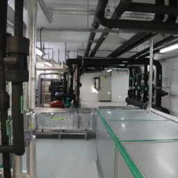 Rozbudowana instalacja rur w kotłowni w Łagiewnikach, czarne i srebrne rury z izolacją, widoczne pompy i skrzynki elektryczne.