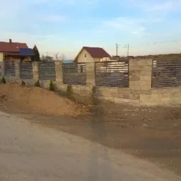 Nowoczesne ogrodzenie panelowe z betonowych bloczków, świeżo posadzone małe tuje wzdłuż ogrodzenia, widoczna pryzma ziemi.