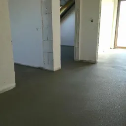 Świeżo wylana, gładka posadzka w surowym wnętrzu budynku, widoczne białe ściany w stanie deweloperskim i otwory drzwiowe.