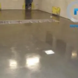 Błyszcząca, szara posadzka przemysłowa w hali. Widoczne żółte zabezpieczenia przy ścianie i metalowa listwa dylatacyjna. Gładka powierzchnia odbija światło.