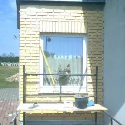 Żółta cegła klinkierowa na elewacji budynku w trakcie prac. Widoczne narzędzia, poziomica i wiadro. Zastosowano listwy dystansowe. Okno z kwiatami w tle.