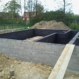 Fundamenty domu z bloczków betonowych, zabezpieczone czarną izolacją, widok z poziomu gruntu, w tle ziemia i zieleń.