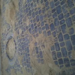 Ułożona kostka brukowa w szarym odcieniu, częściowo zasypana piaskiem, tworząca dekoracyjny wzór z okrągłym elementem w centrum.