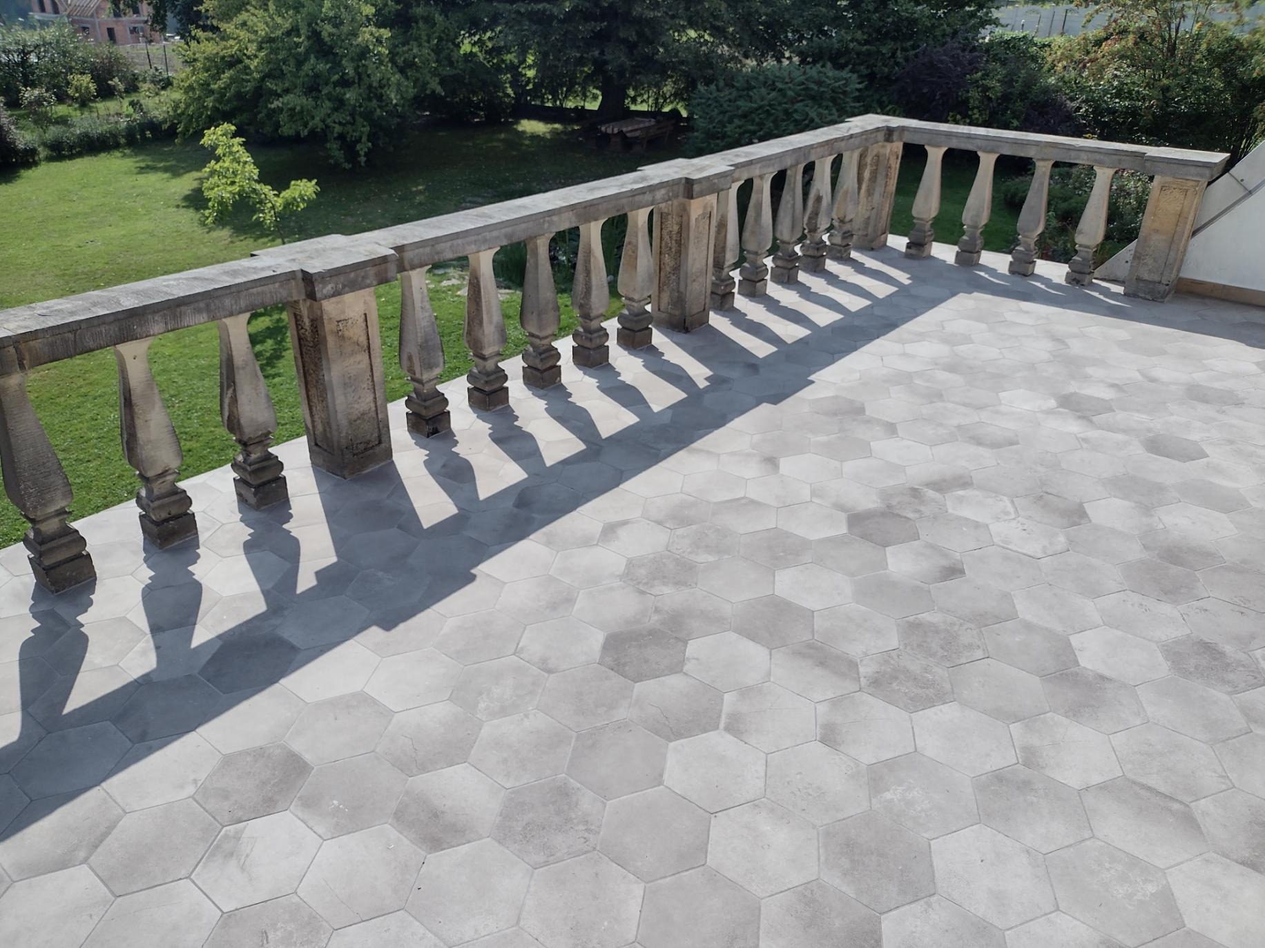 Balkon z szarą, heksagonalną podłogą, kamienna balustrada rzucająca cień, widok na zielony ogród.
