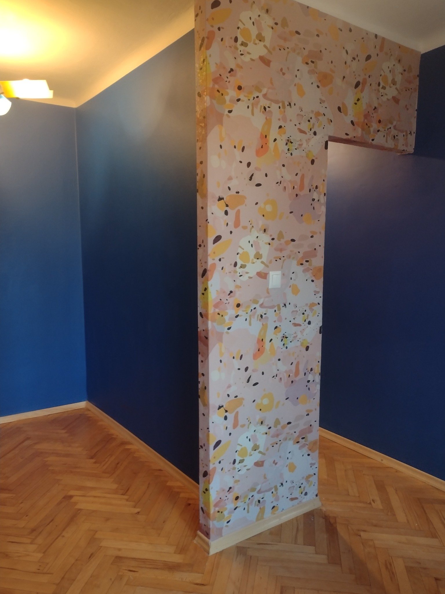 Pomieszczenie z ciemnoniebieskimi ścianami, parkietem w jodełkę i ścianą akcentową z wzorem terrazzo, widoczny włącznik światła.