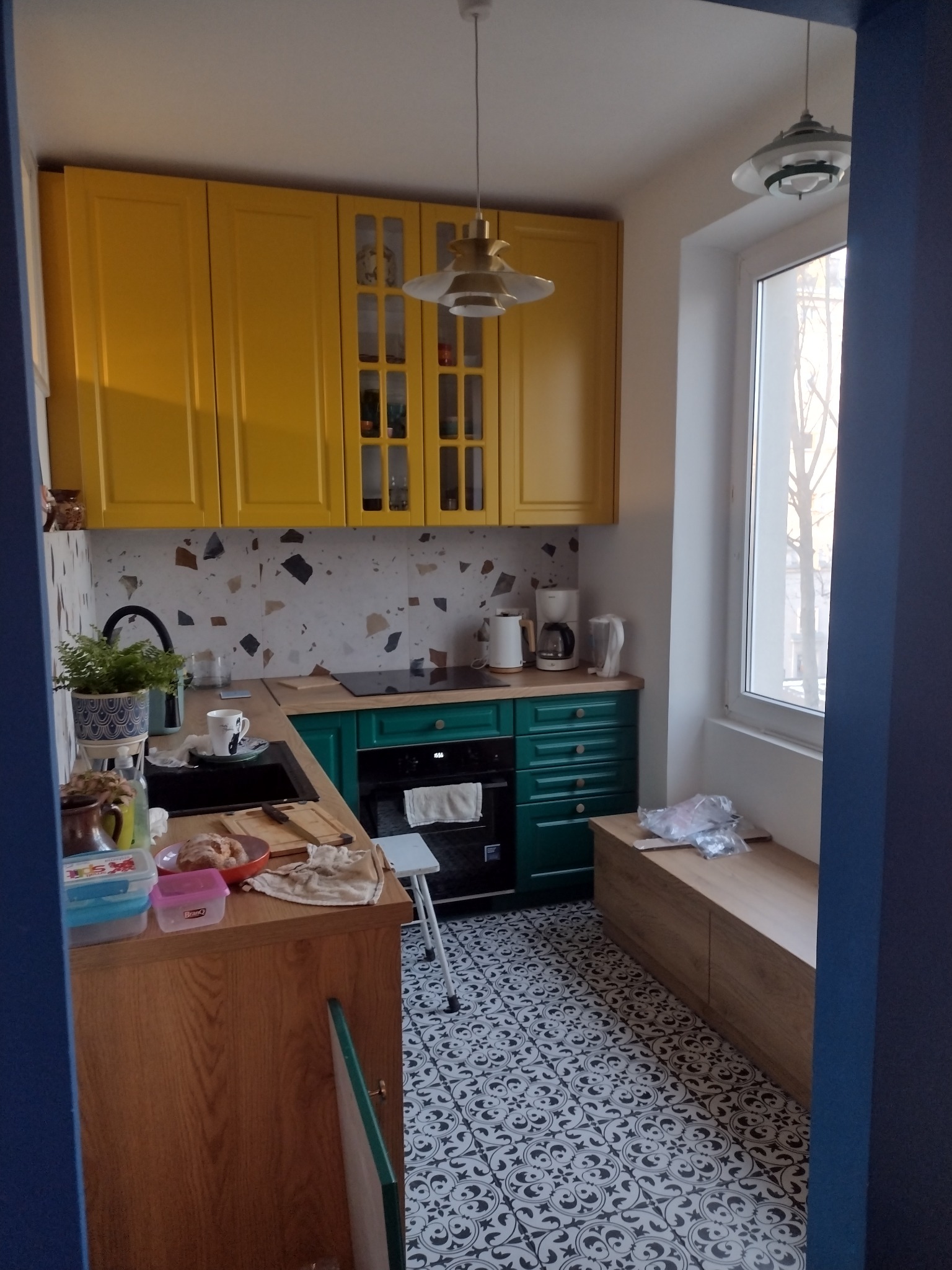 Kuchnia po remoncie z żółtymi szafkami górnymi, zielonymi dolnymi, wzorzystą mozaiką na ścianie i dekoracyjną podłogą w stylu retro.