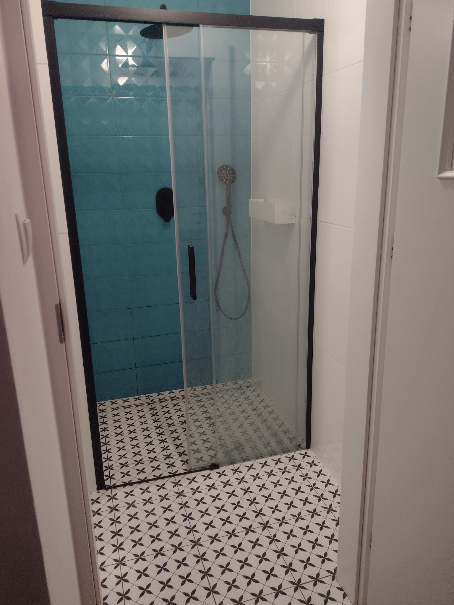 Nowoczesna kabina prysznicowa z czarną ramą i turkusowymi płytkami w tle, widoczna przez otwarte drzwi łazienki z biało-czarną mozaiką podłogową.