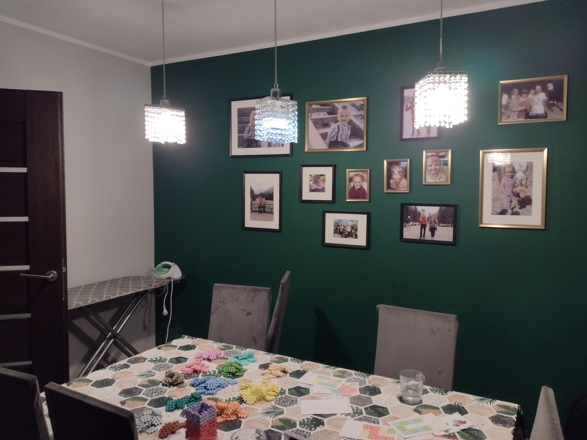 Zielona ściana z galerią rodzinnych zdjęć w ramkach, oświetlona trzema kryształowymi lampami wiszącymi, stół z rozłożonymi klockami do układania i krzesłami, deska do prasowania z żelazkiem w tle.