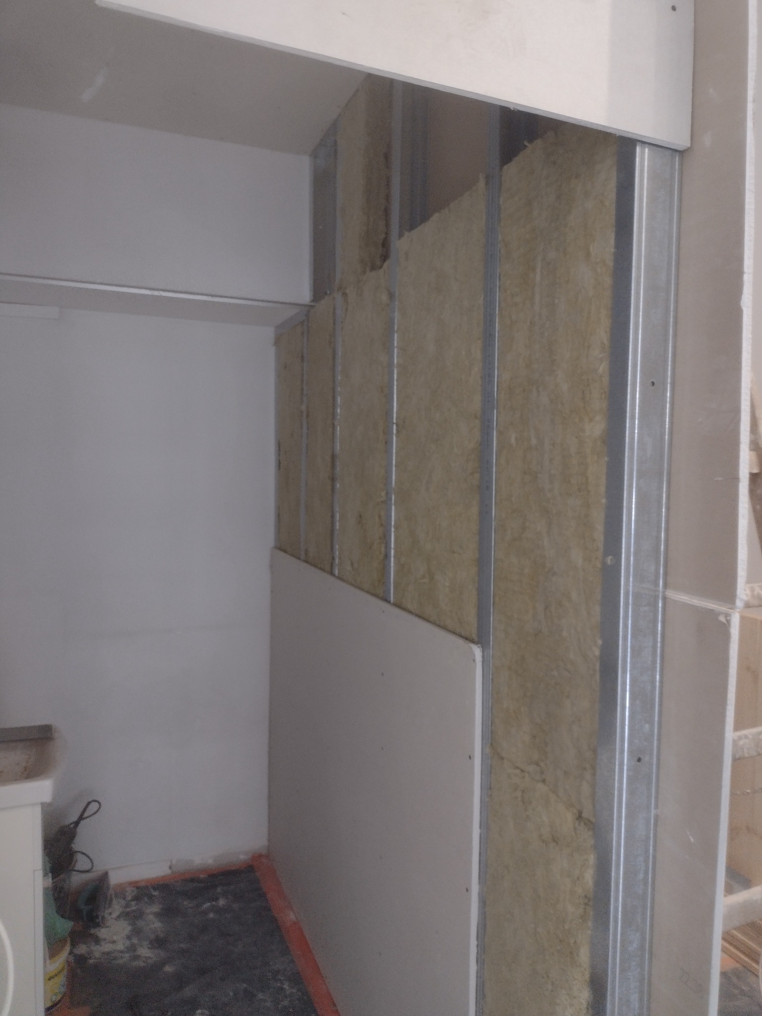 Wnętrze w trakcie prac: metalowy stelaż z wełną mineralną, częściowo zakryty płytą gipsowo-kartonową, widoczne elementy instalacji w tle.